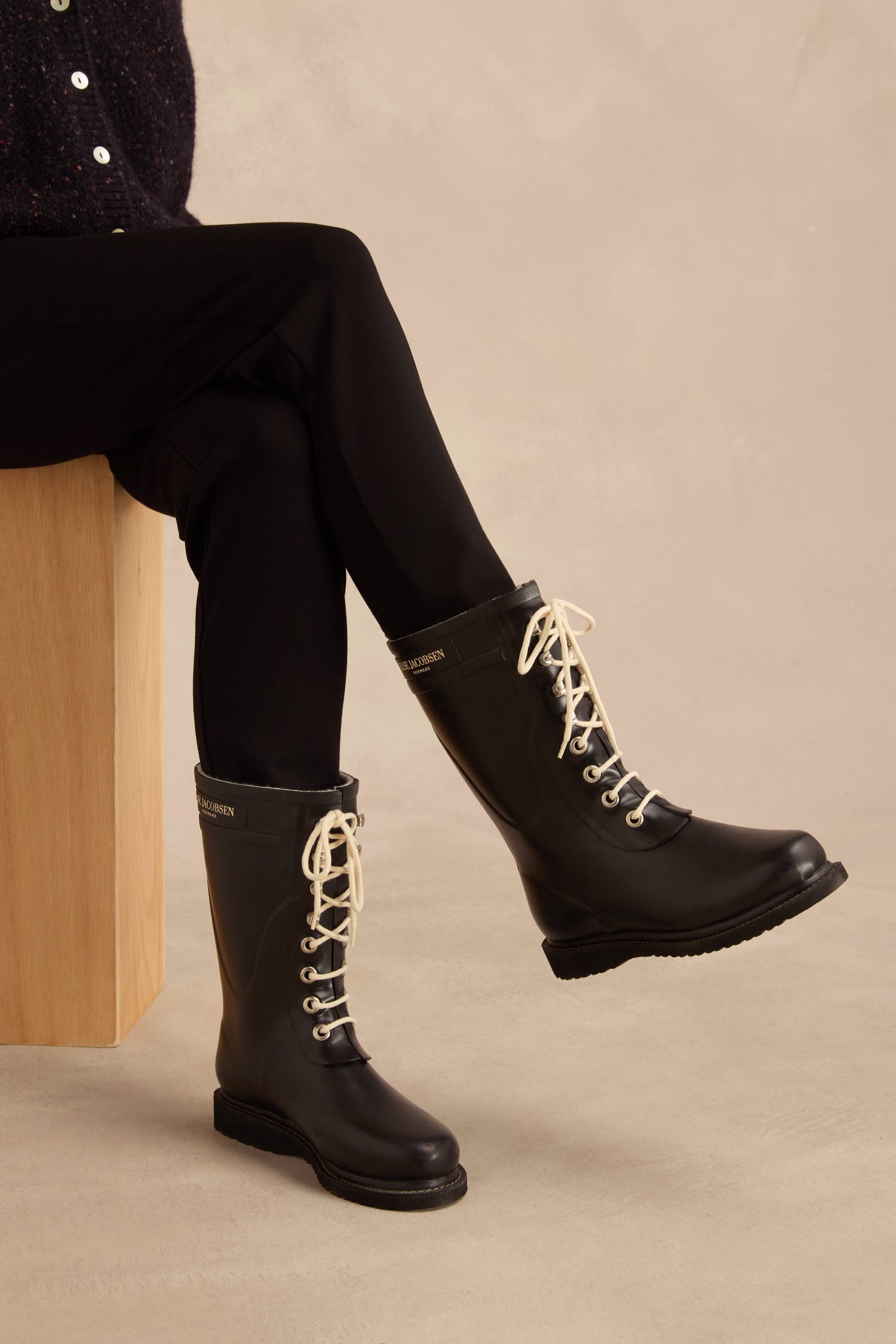 A person seated on a wooden block, dressed in black pants and the Lace Up Wellington Boots—crafted from natural rubber for stylish, reliable wet-weather protection—set against a neutral background.