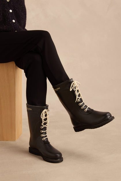 A person seated on a wooden block, dressed in black pants and the Lace Up Wellington Boots—crafted from natural rubber for stylish, reliable wet-weather protection—set against a neutral background.