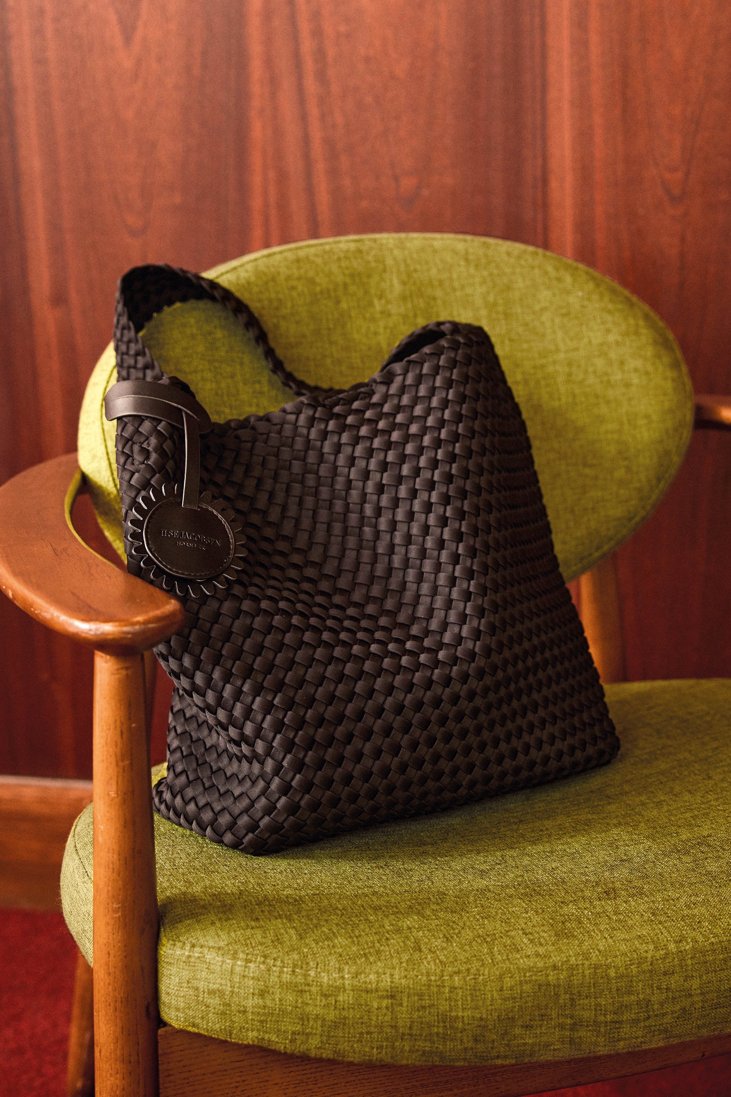 A Black Braided Shoulder Bag, style IJSHELLBAG04, featuring a round tag, rests on a vintage green upholstered chair with wooden arms, set against a wood-paneled wall.