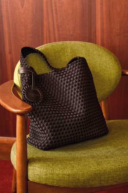 A Black Braided Shoulder Bag, style IJSHELLBAG04, featuring a round tag, rests on a vintage green upholstered chair with wooden arms, set against a wood-paneled wall.