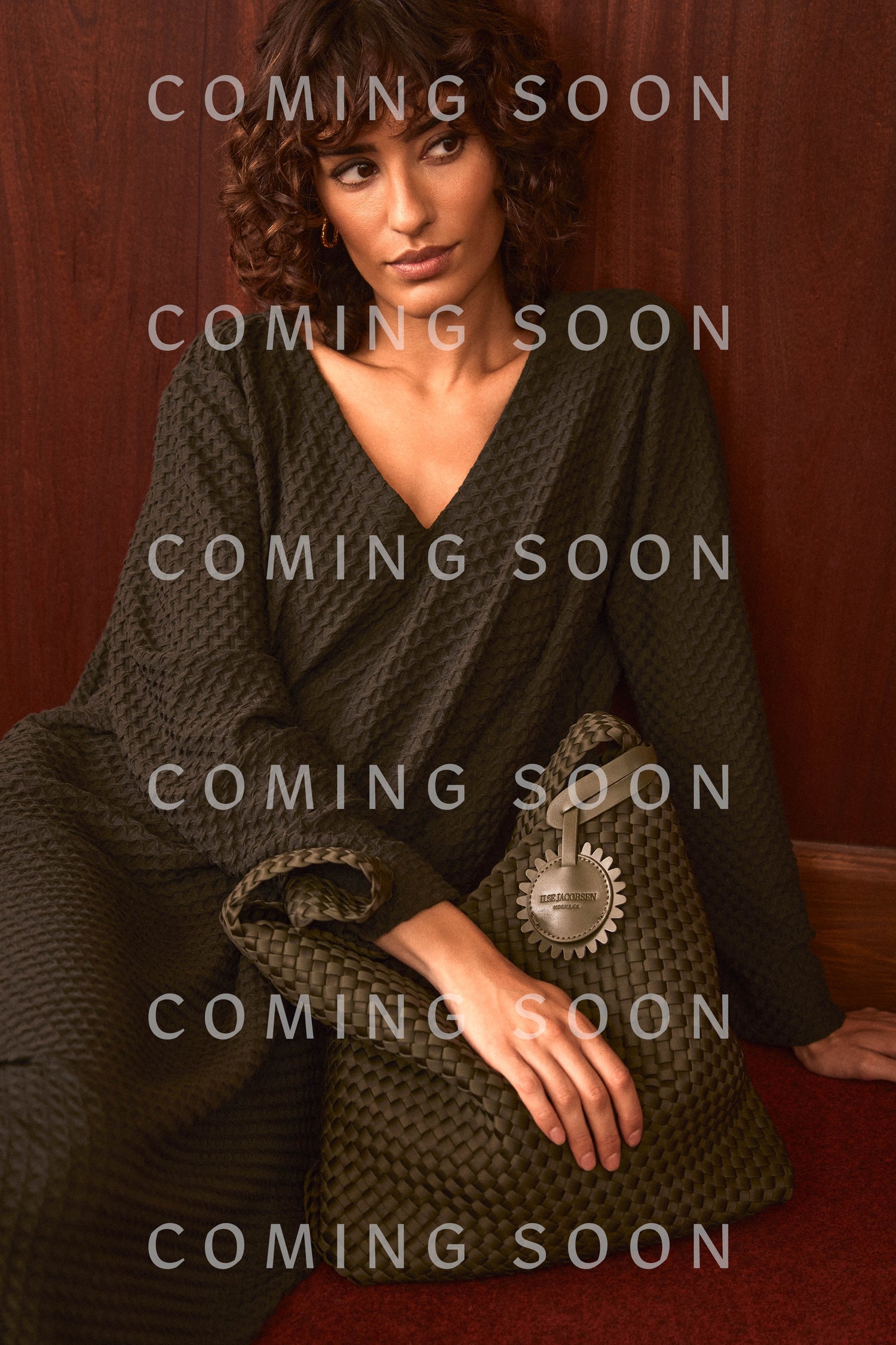 A woman in a textured dark dress sits against a wooden wall with a large, woven Olive Braided Shoulder Bag; “COMING SOON” text overlays the image repeatedly.