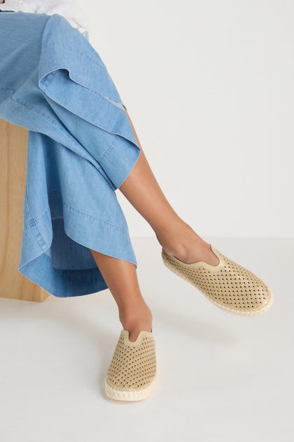 A person sits on a wooden chair with legs crossed, wearing light blue wide-leg pants and Tulip Glitter Slip On Shoes in beige by Ilse Jacobsen, featuring perforated detailing.