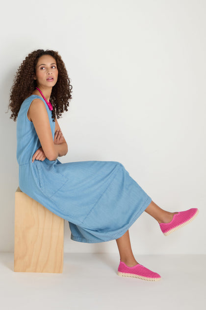 A woman with curly hair sits on a wooden block against a plain white background, wearing a sleeveless denim dress, pink necklace, and Rose Tulip Slip on Shoes known for their lightweight comfort.