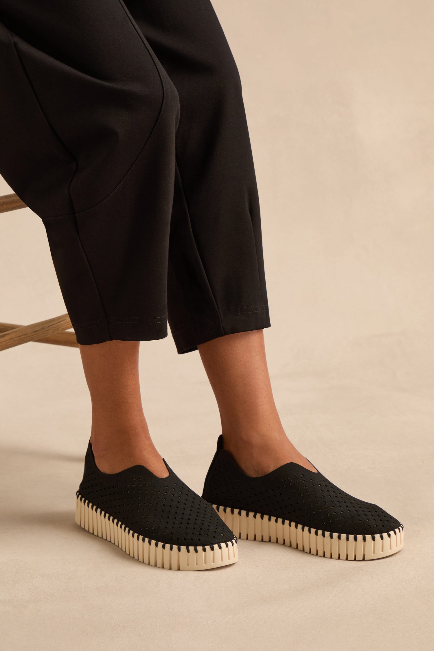 A person is seated on a chair, wearing the Tulip Platform Slip On Shoes in black—crafted from recycled microfiber with thick white soles—paired with black ankle-length pants, set against a neutral background.