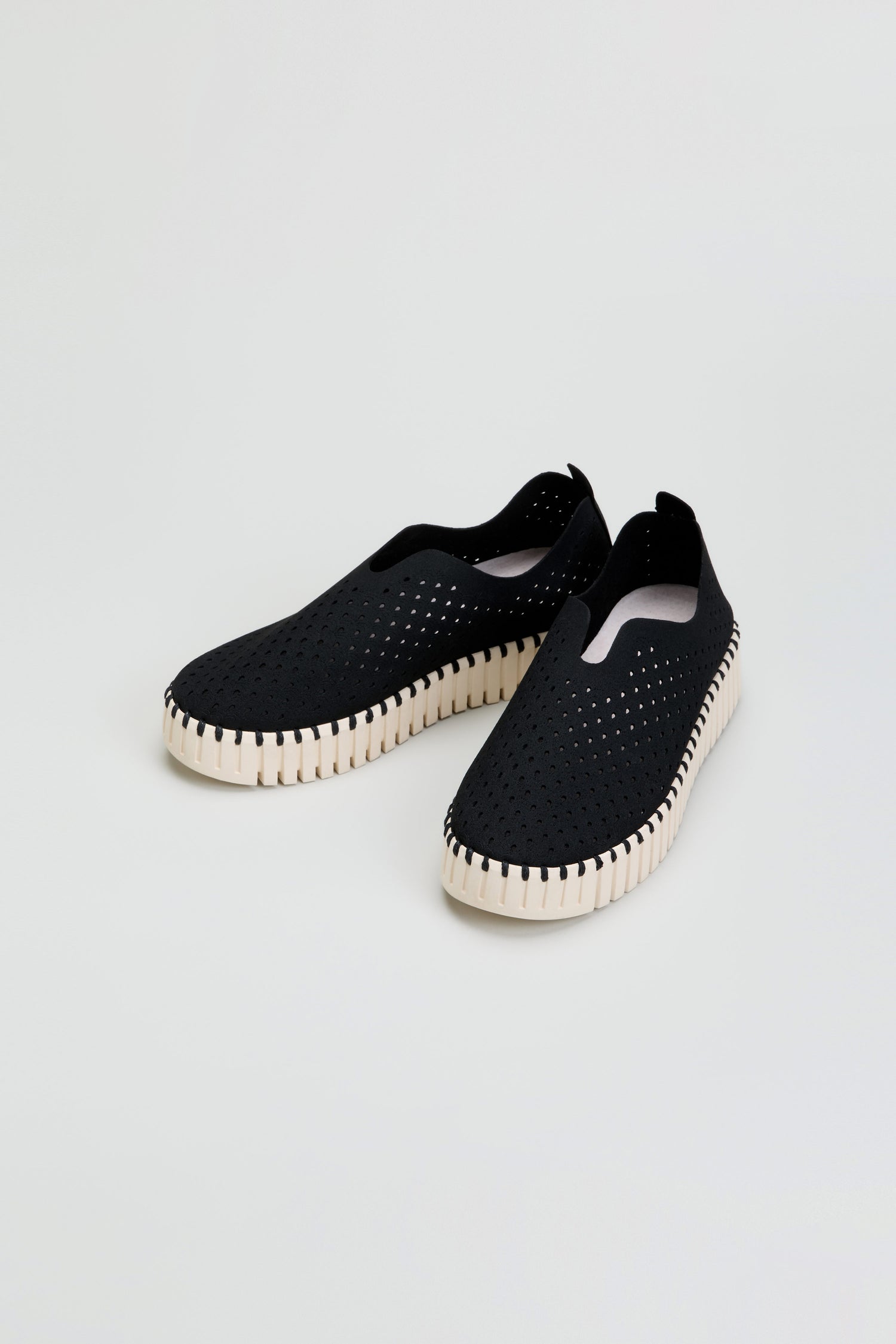 A pair of black Tulip Platform Slip On Shoes with perforated uppers and white textured soles, displayed on a plain light background. These breathable slip-on shoes are crafted from recycled microfiber, combining eco-friendly materials with effortless style.