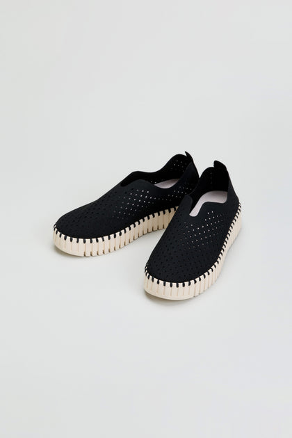 A pair of black Tulip Platform Slip On Shoes with perforated uppers and white textured soles, displayed on a plain light background. These breathable slip-on shoes are crafted from recycled microfiber, combining eco-friendly materials with effortless style.