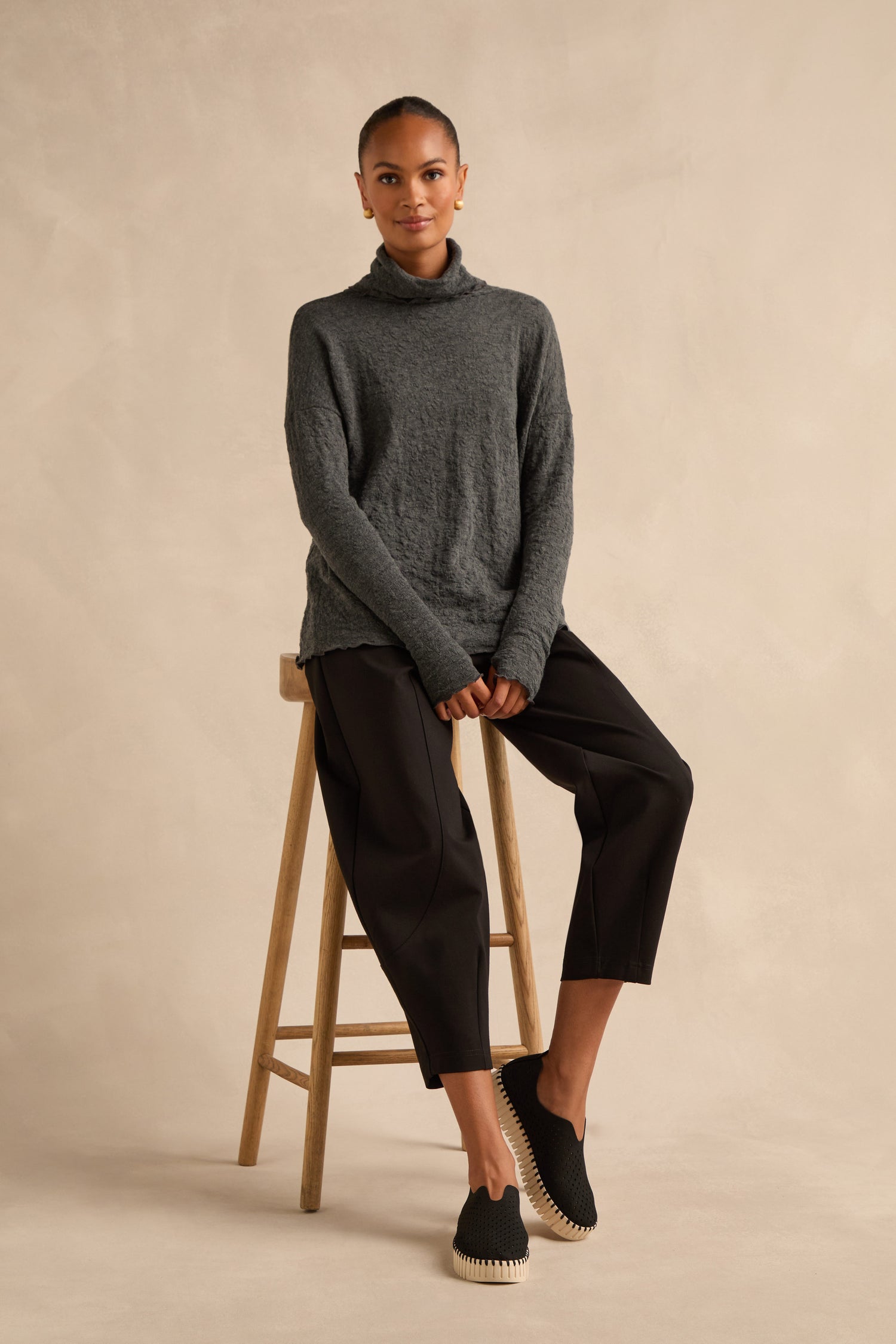 A person sits on a wooden stool wearing a dark gray turtleneck sweater, black cropped pants, and breathable slip-on shoes against a beige background.