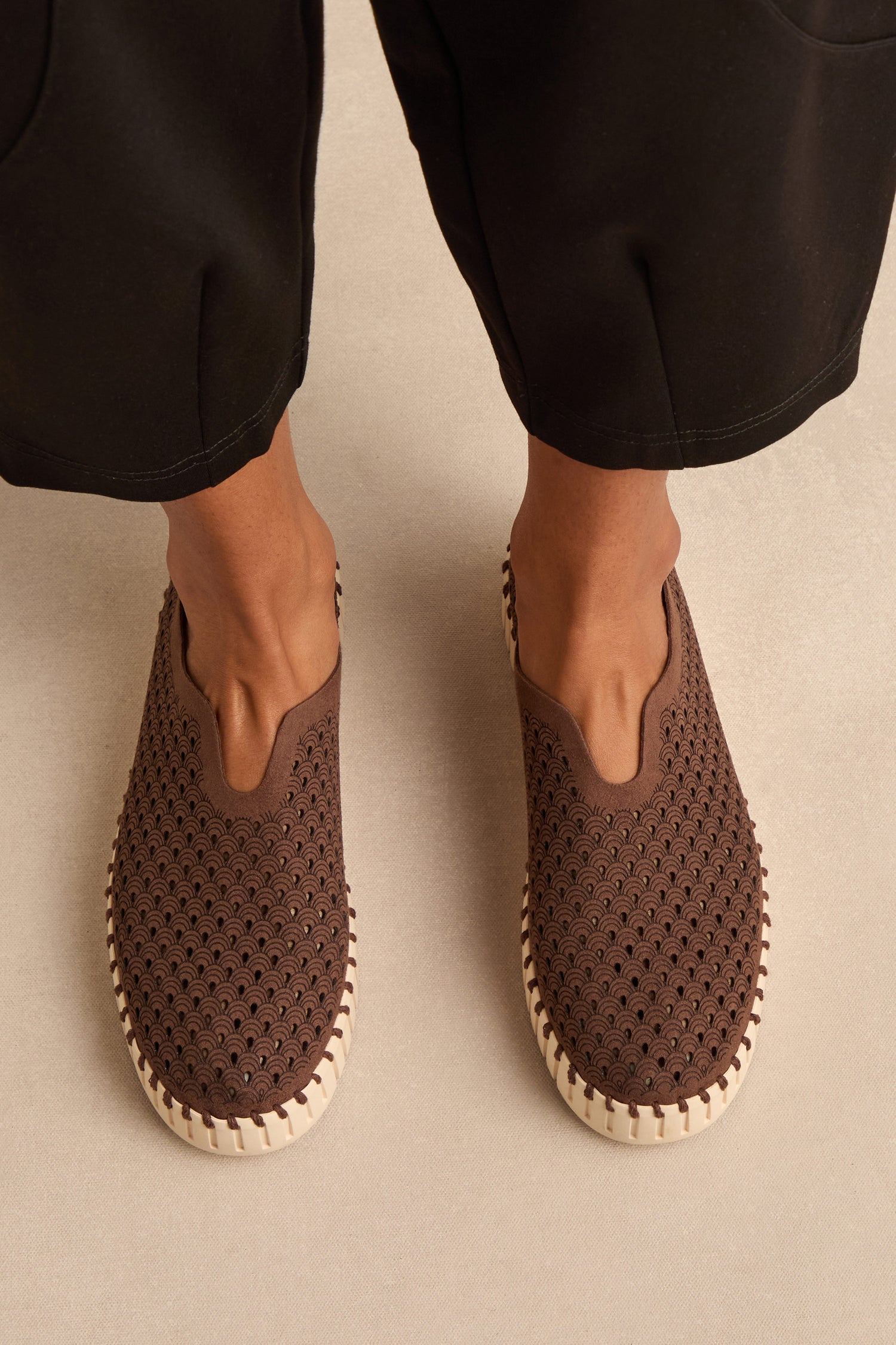A person wears Tulip Platform Slip On Shoes with a brown, perforated upper and white soles, standing on a light background. Only their lower legs and feet are shown, with black pants cropped above the ankles—ideal for highlighting these eco-friendly slip-ons made from recycled microfiber.