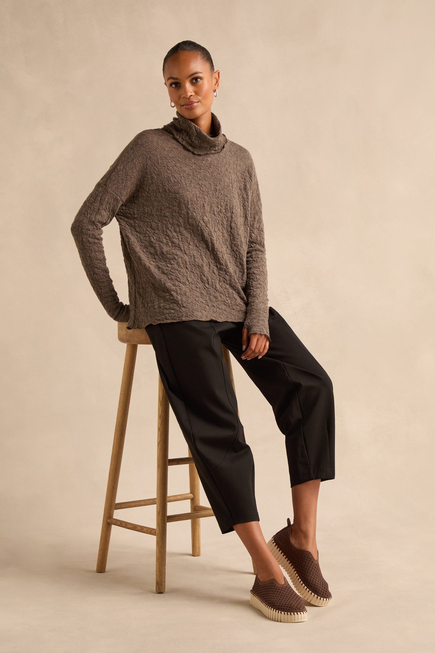 A woman with her hair pulled back sits on a wooden stool wearing a textured brown turtleneck sweater, black cropped trousers, and Tulip Platform Slip On Shoes made from recycled microfiber, against a beige backdrop.