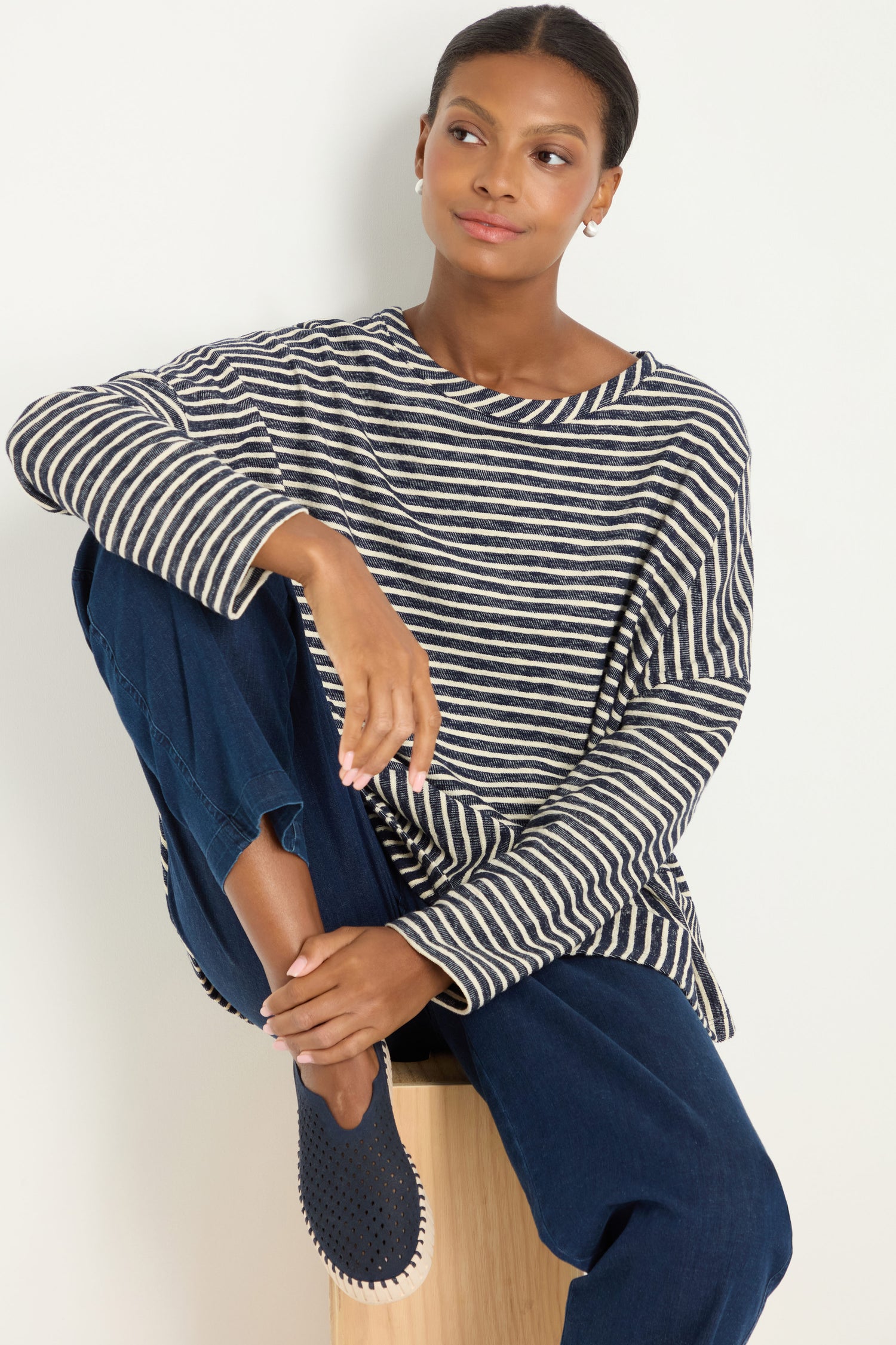 A woman wearing a black and white striped long-sleeve shirt, navy pants, and Tulip Platform Slip on Shoes sits on a wooden stool with one knee raised, looking to the side.