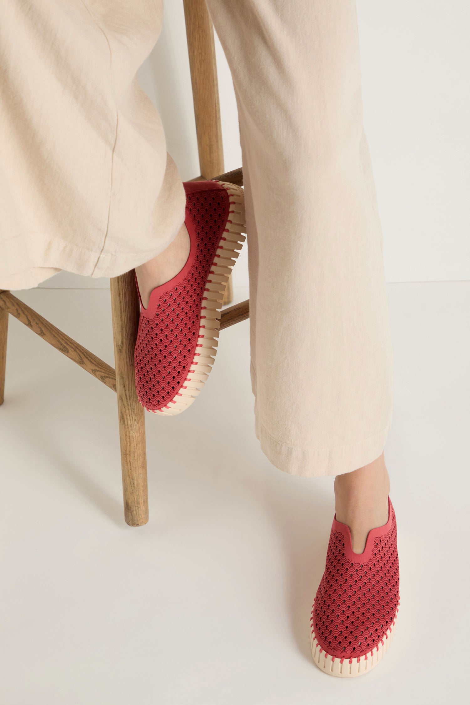 A person sits on a wooden stool wearing light beige pants and red Tulip Platform Slip On Shoes by Ilse Jacobsen, featuring perforated recycled microfibre uppers and light-colored soles.