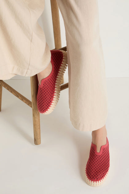 A person sits on a wooden stool wearing light beige pants and red Tulip Platform Slip On Shoes by Ilse Jacobsen, featuring perforated recycled microfibre uppers and light-colored soles.