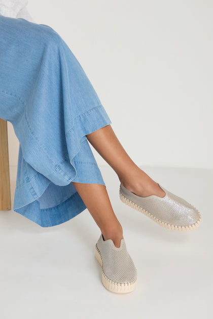 A person sits cross-legged in a light blue denim skirt, wearing Tulip Glimmer Platform Slip on Shoes with textured soles, set against a plain light background.