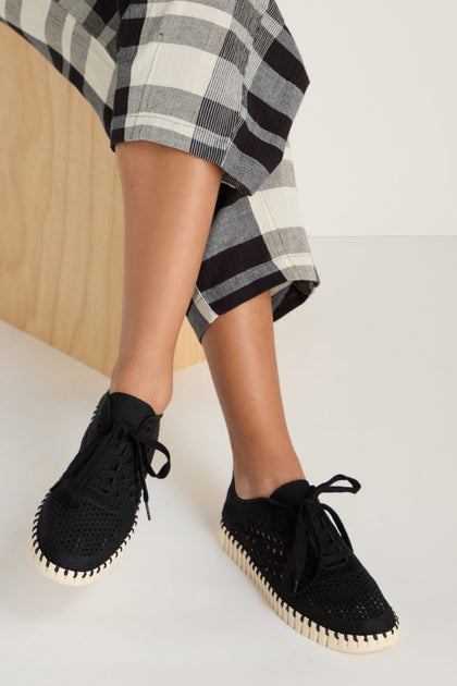 A person wearing Tulip Embroidered Lace Up Trainers with white soles and black-and-white plaid pants, seated on a light wooden bench.