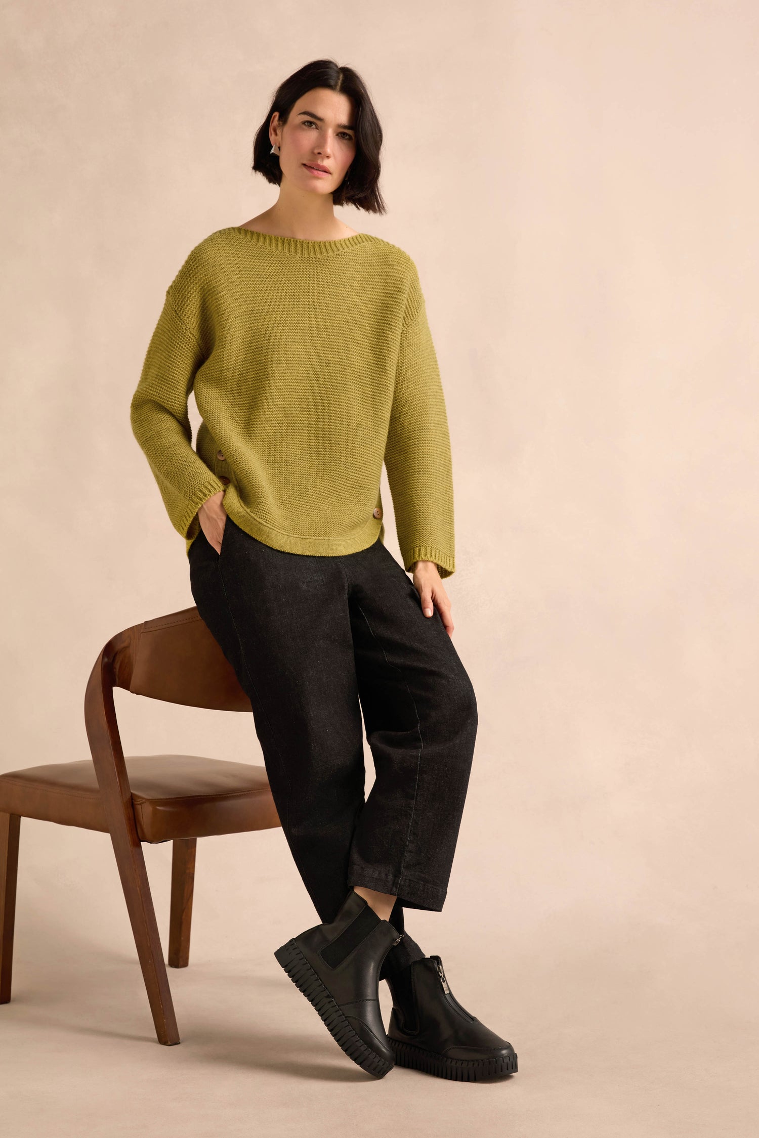 A person with short dark hair wearing a loose green sweater, black pants, and Tulip Zip Chelsea Boots made from recycled microfibre sits on a wooden chair against a plain beige background.