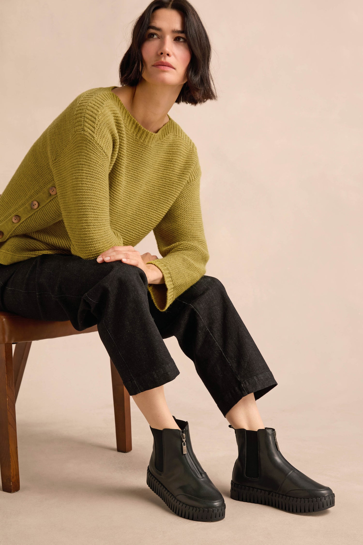 Woman with short dark hair sits on a wooden chair, wearing a green sweater, black pants, and stylish Tulip Zip Chelsea Boots crafted from recycled microfibre with a cozy faux shearling lining, against a neutral background.