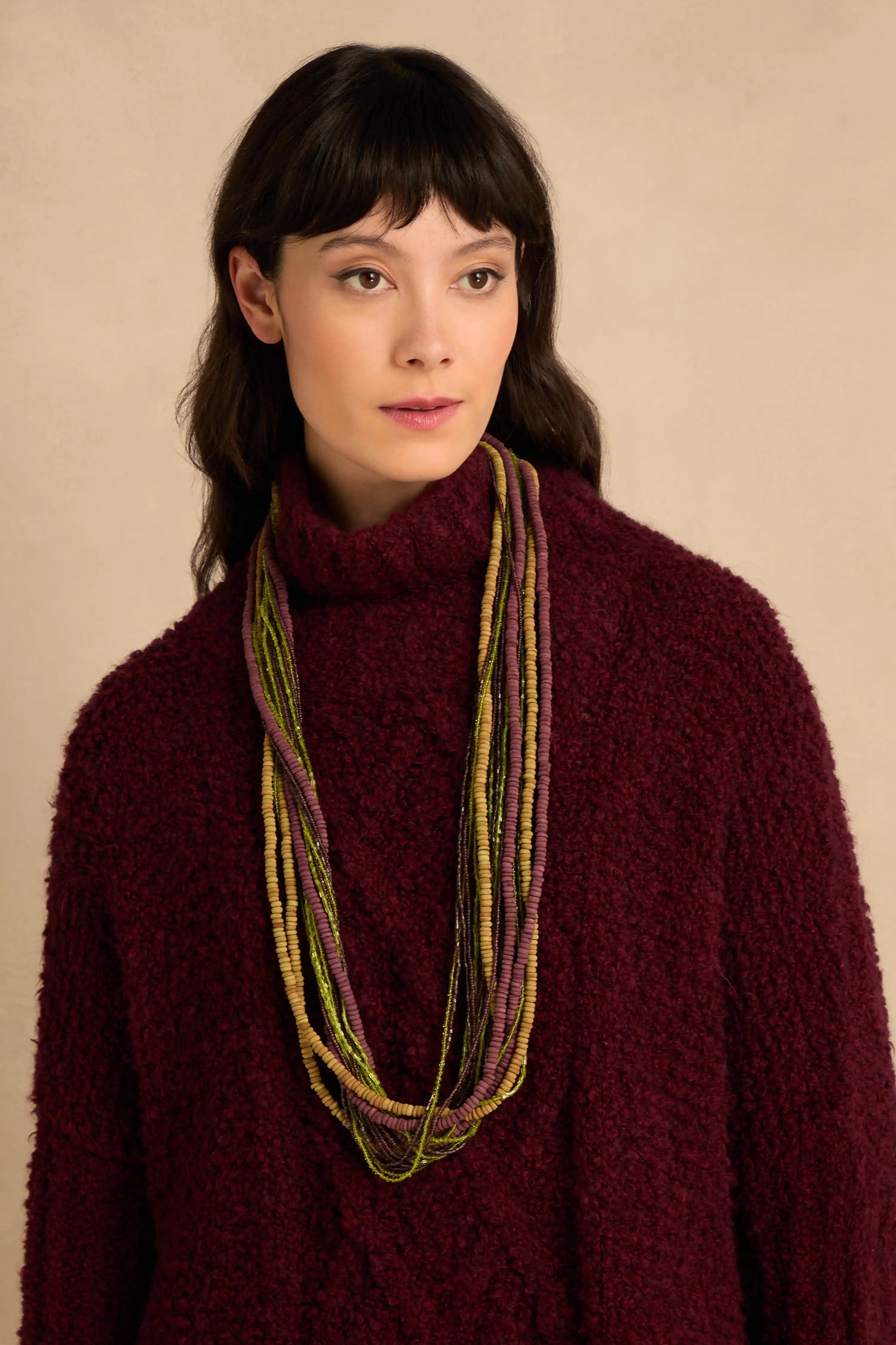 A Mango Wood & Bead Waterfall Necklace featuring multi-strand green and purple beads worn over a dark red turtleneck sweater.