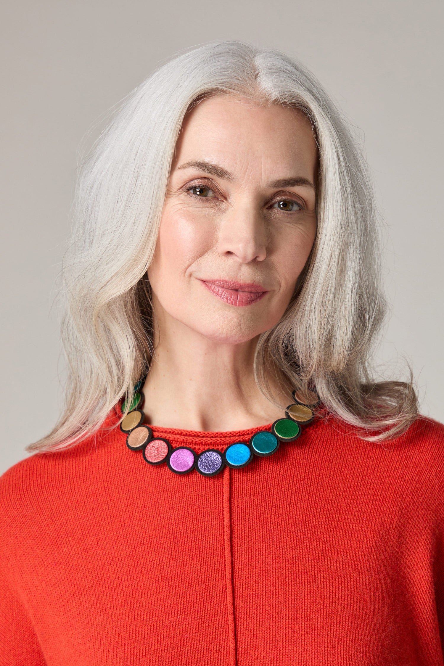 A woman with grey hair wearing a red sweater accessorized with a Metallic Rainbow Leather Disc Necklace.