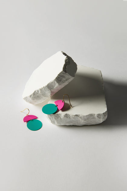 The Colour Pop Circle Drop Earrings bring statement jewelry to life, showcasing vibrant circular pink and turquoise shapes against a white stone backdrop, with one earring elegantly hanging on the stone.