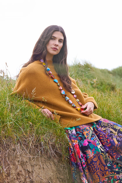 A woman with long dark hair sits outdoors on a grassy slope, wearing a mustard sweater, a vibrant floral skirt, and the Long Kandinsky Pebble Resin Necklace.