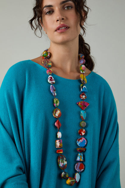 A woman wears a loose turquoise top and the Long Kandinsky Pebble Resin Necklace, standing against a plain light background.