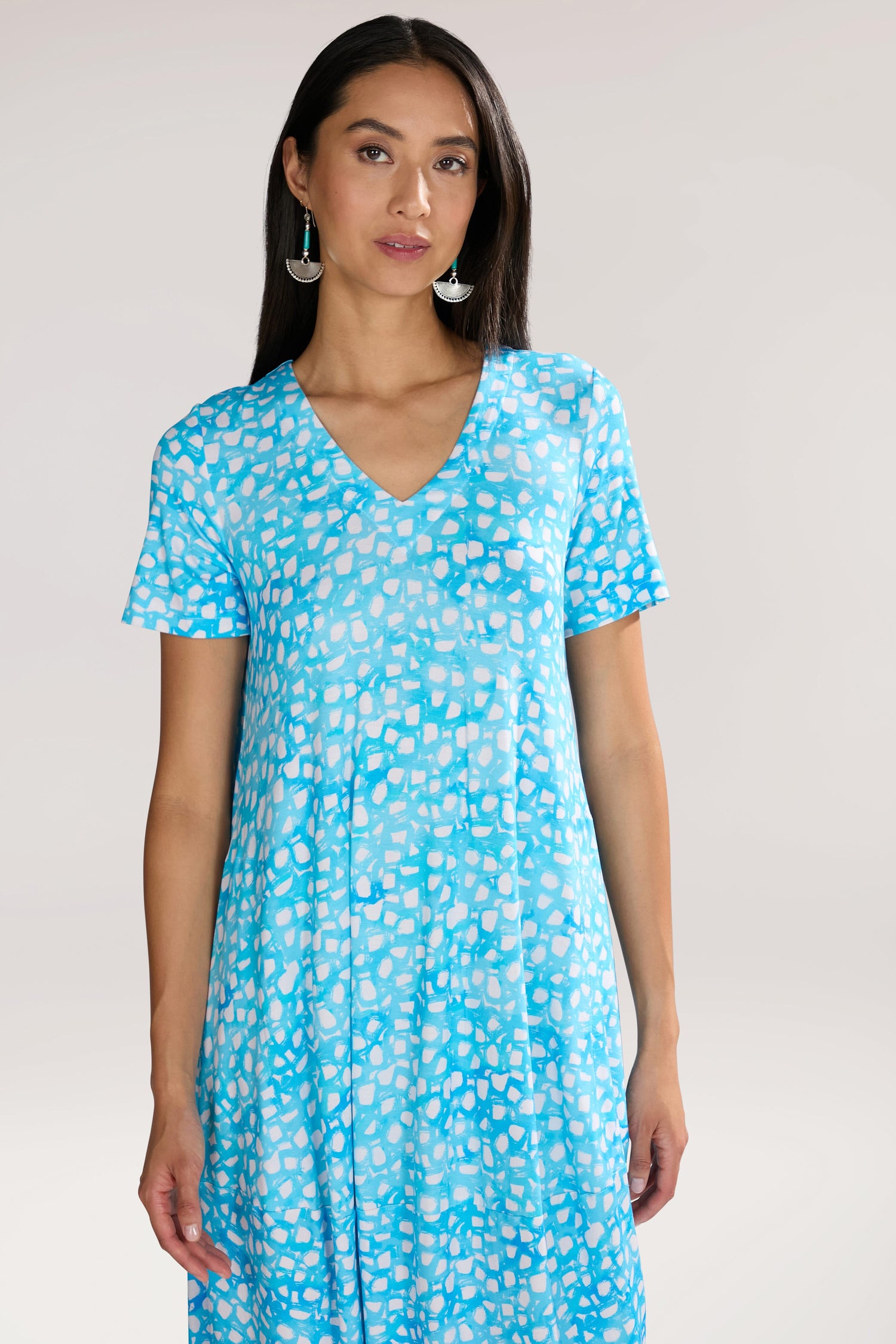 A woman with long dark hair wearing a blue and white Pebble Jersey Print Bubble Dress, looking at the camera with a neutral expression. The short-sleeved dress features an abstract bubble motif.