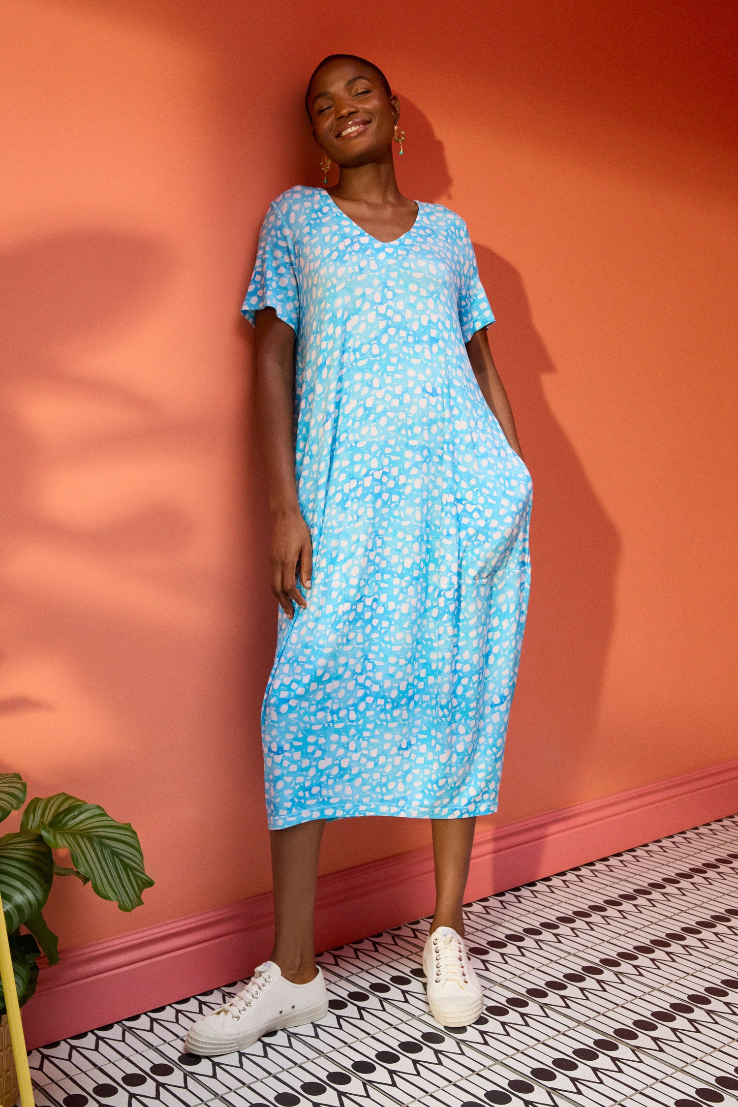 A person wearing a Pebble Jersey Print Bubble Dress and white sneakers stands against a coral-colored wall, with a plant nearby.