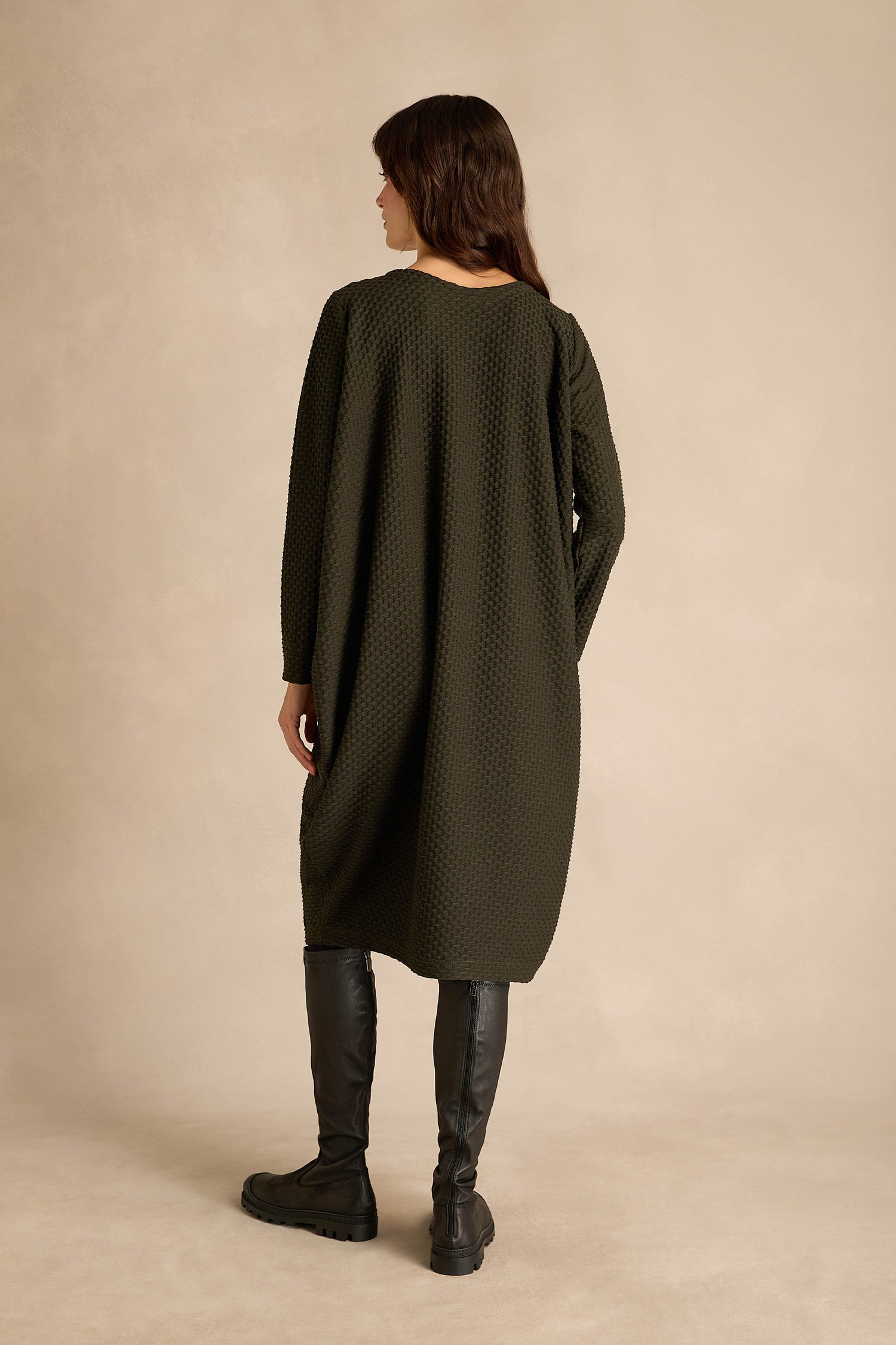 A person stands with their back to the camera, wearing a pine green Waffle Textured Jersey Drape Dress and black knee-high boots against a beige background.