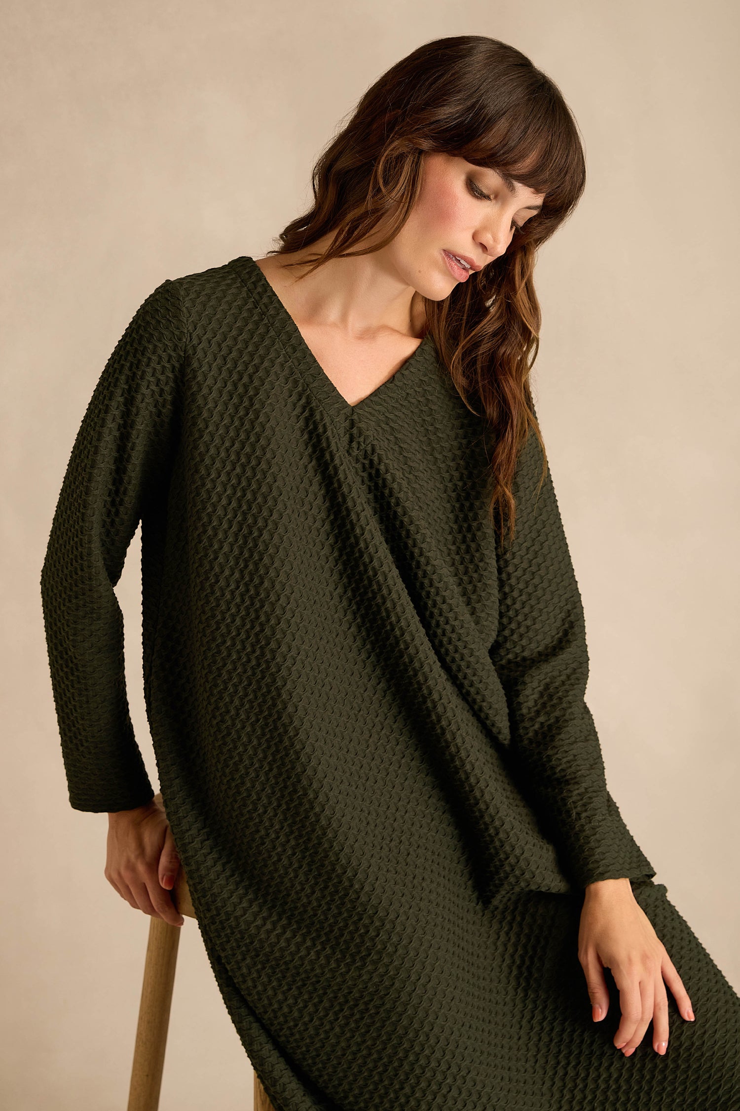 A woman sits on a stool against a neutral background, looking down with her hand resting on her lap, wearing the Waffle Textured Jersey Drape Dress in pine green with long sleeves.