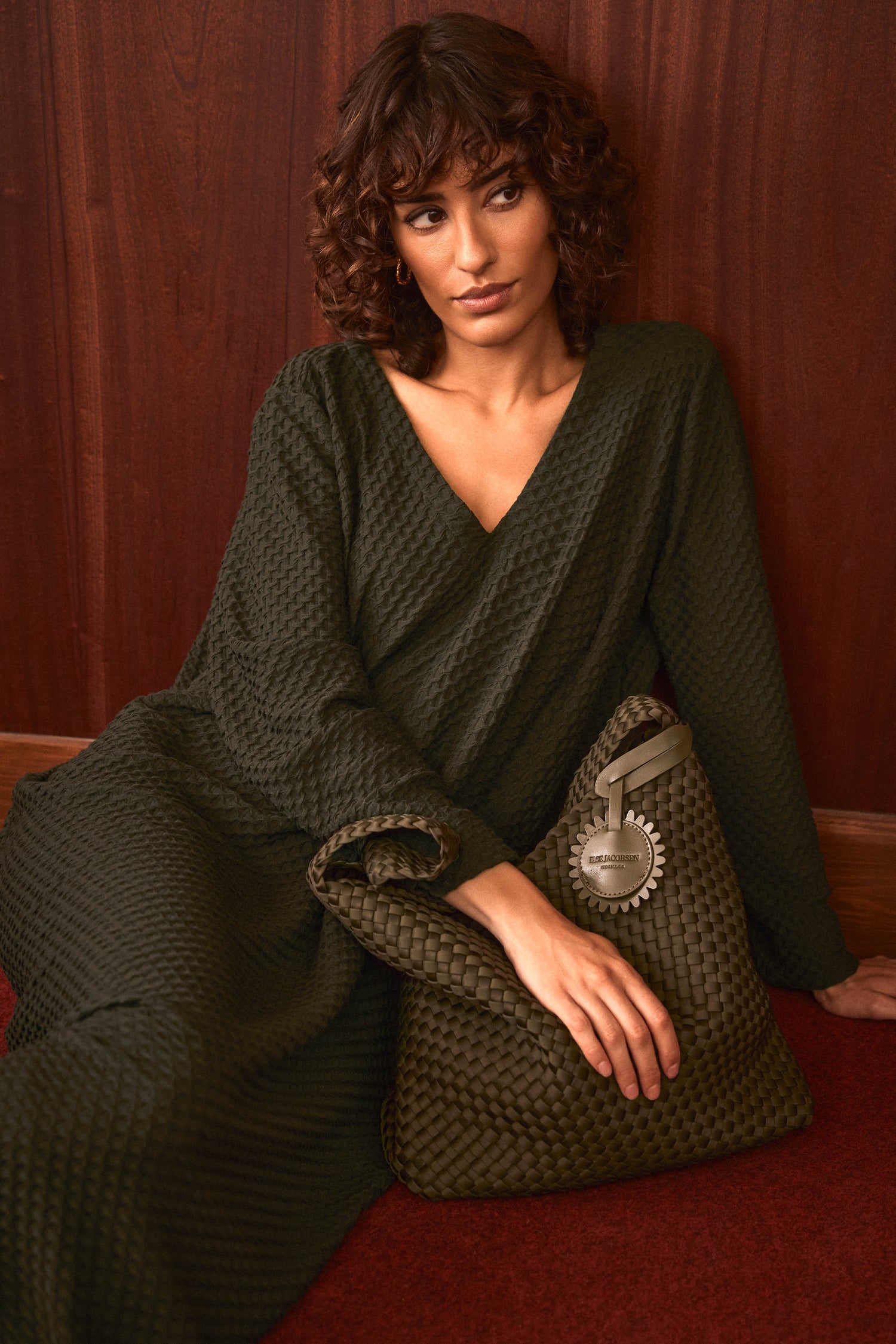 Woman with curly hair wearing the Waffle Textured Jersey Drape Dress sits on the floor against a wood-paneled wall, holding a large woven green handbag.