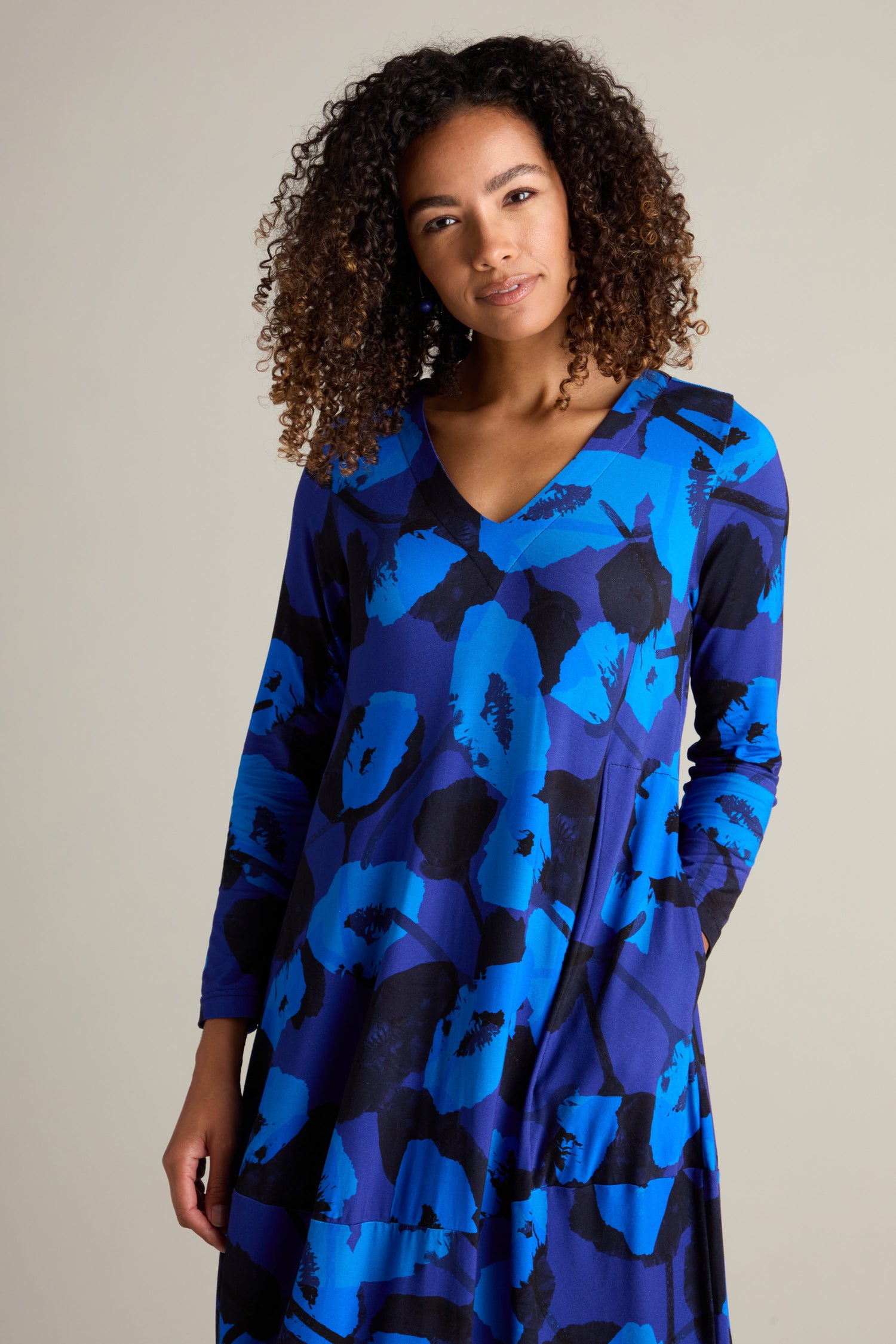 A person with curly hair is wearing a Flowering Poppies Bubble Dress, featuring a blue patterned long-sleeve design with a contemporary silhouette, standing against a plain background.