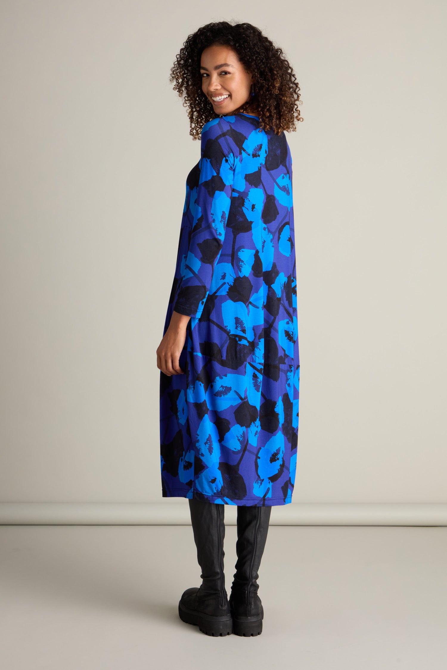 A woman stands facing slightly backward, smiling at the camera. She is wearing the Flowering Poppies Bubble Dress—a blue and black patterned, long-sleeve dress with a blue poppy print—and black knee-high boots. The background is plain and light-colored.