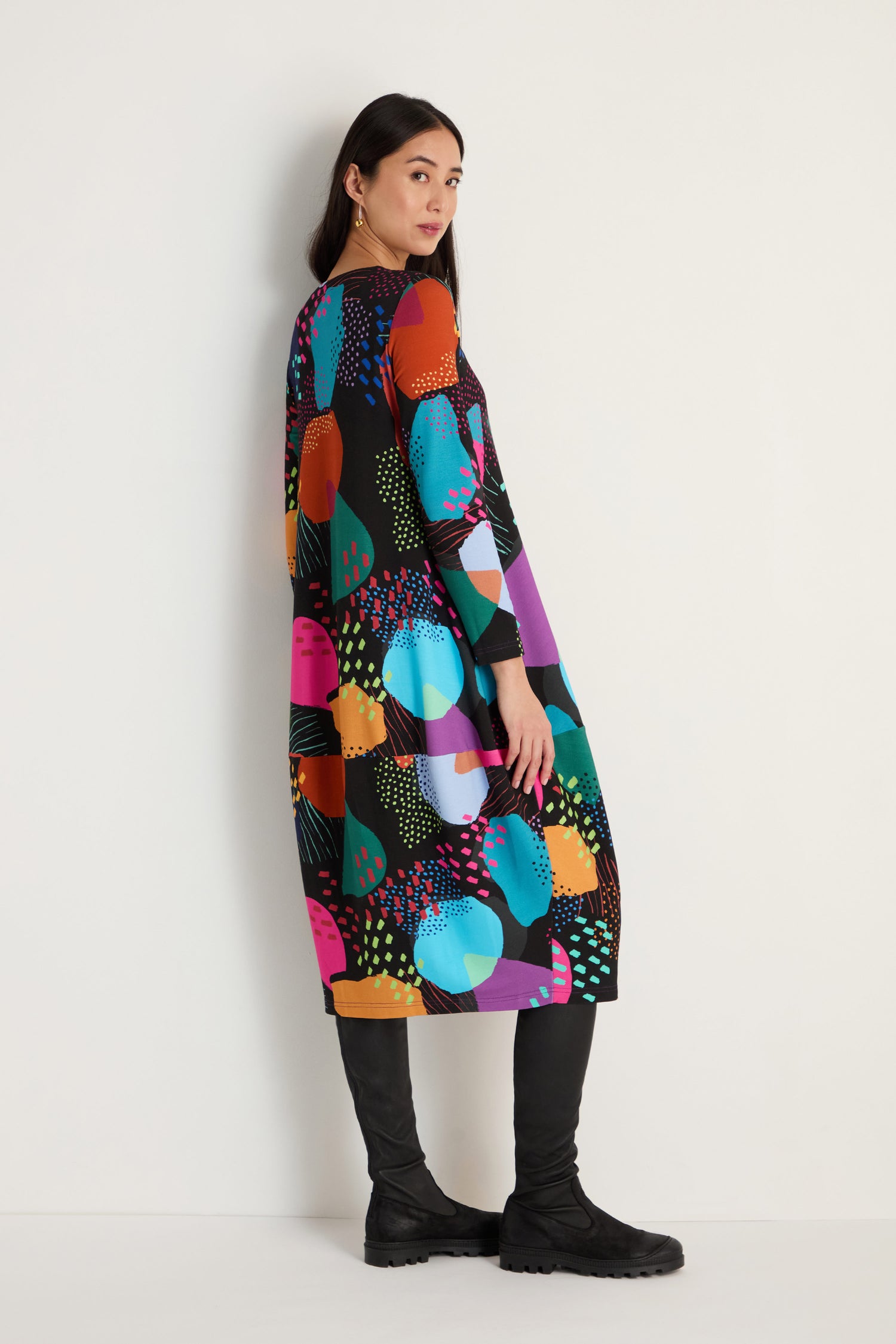 A woman stands against a white wall wearing the Mark Making Print Jersey Bubble Dress with long sleeves and black knee-high boots.