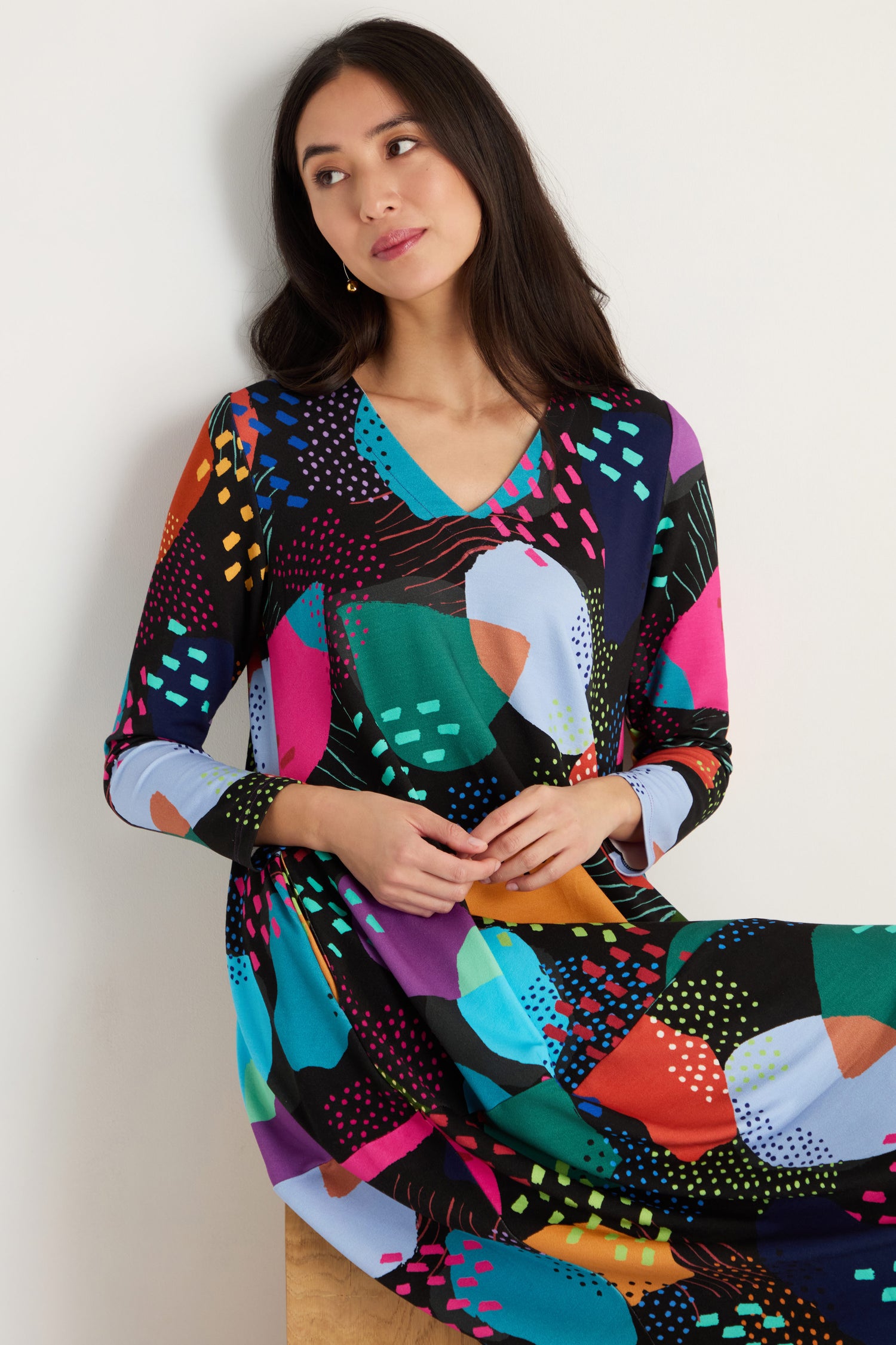A woman with long dark hair wears the Mark Making Print Jersey Bubble Dress, featuring a colorful pattern, long sleeves, and a V-neck, while sitting against a plain light background.