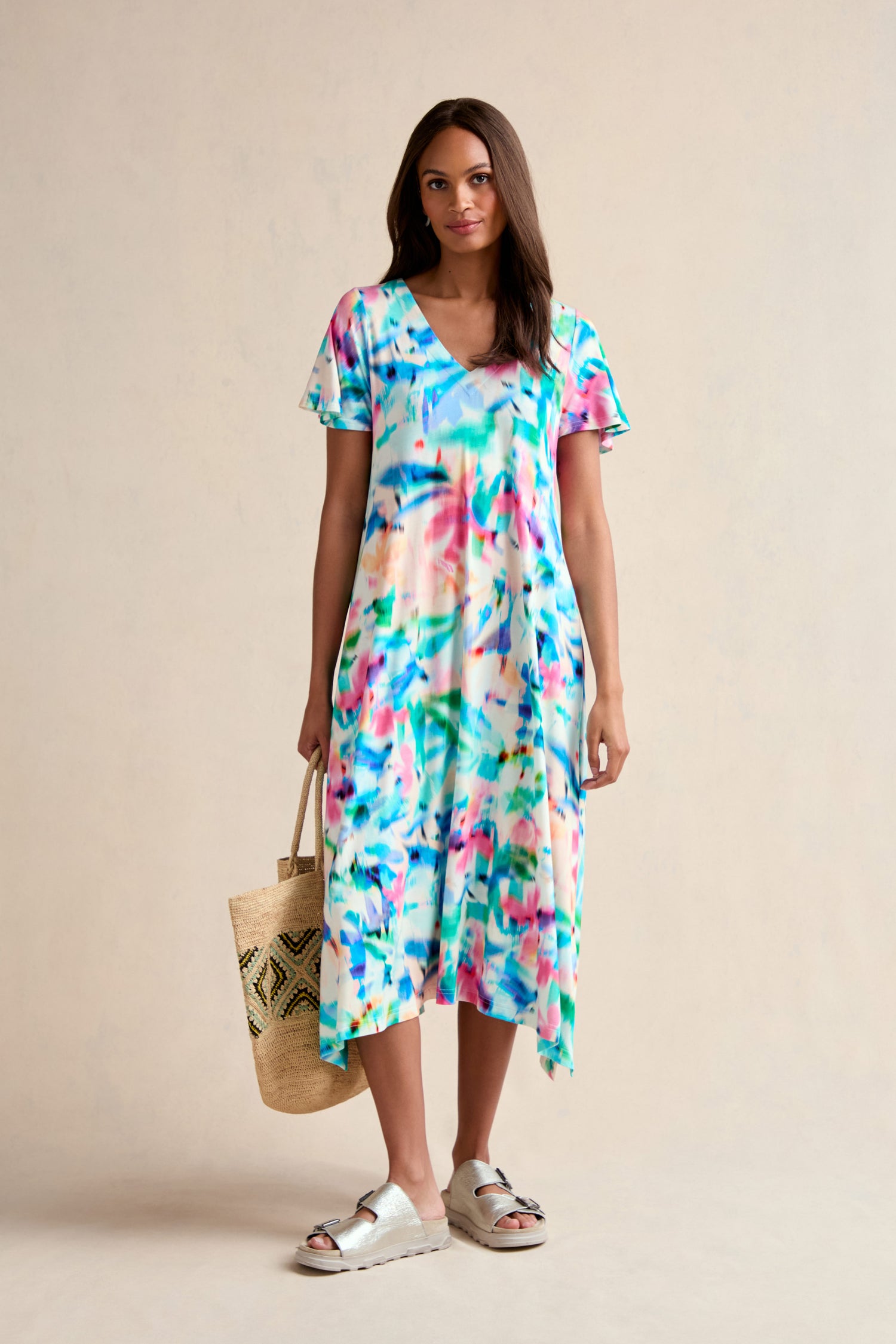 A woman stands facing forward wearing the vibrant Petal Haze Jersey Flare Dress, white sandals, and holding a woven tote bag against a plain beige background.