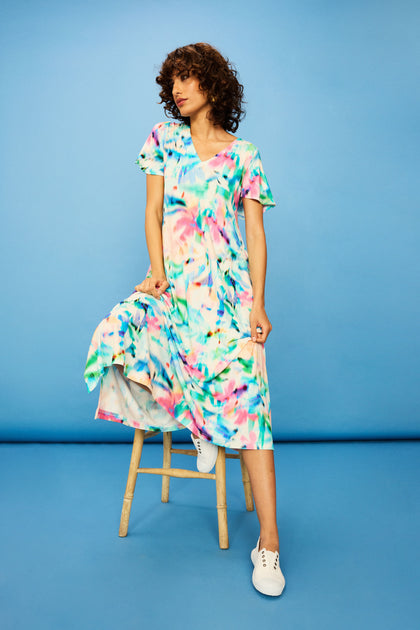 Wearing the Petal Haze Jersey Flare Dress and white sneakers, a person sits on a wooden stool against a plain blue background, holding part of the dress in one hand.