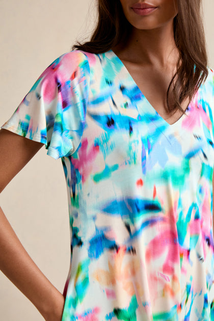 A person wears the Petal Haze Jersey Flare Dress, a short-sleeve V-neck style featuring a colorful abstract pattern in blue, pink, and green.