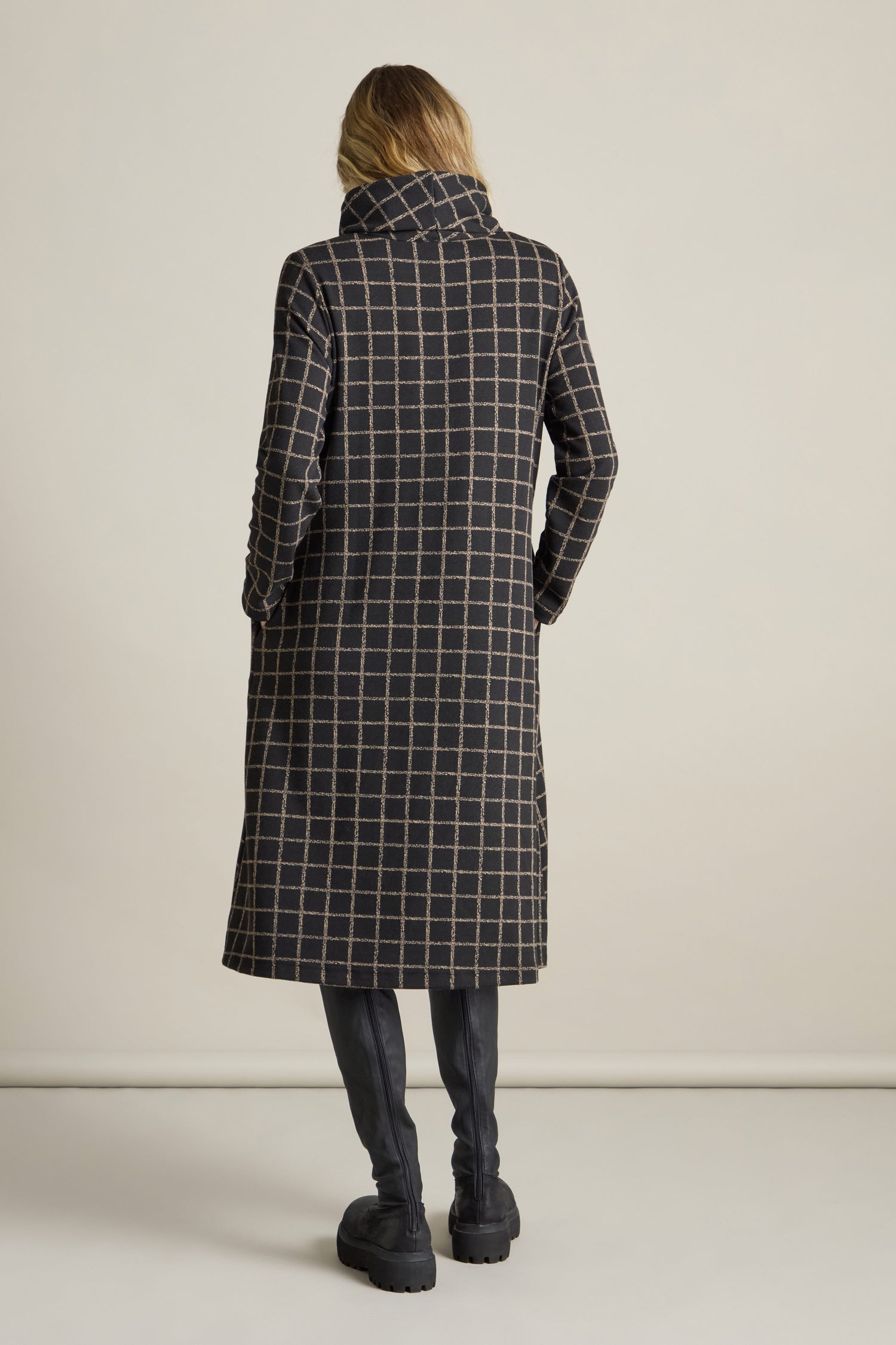 A person stands with their back to the camera, wearing a long plaid coat and black boots against a neutral background, offering a contemporary twist reminiscent of the stylish Broken Check Jersey Dress.