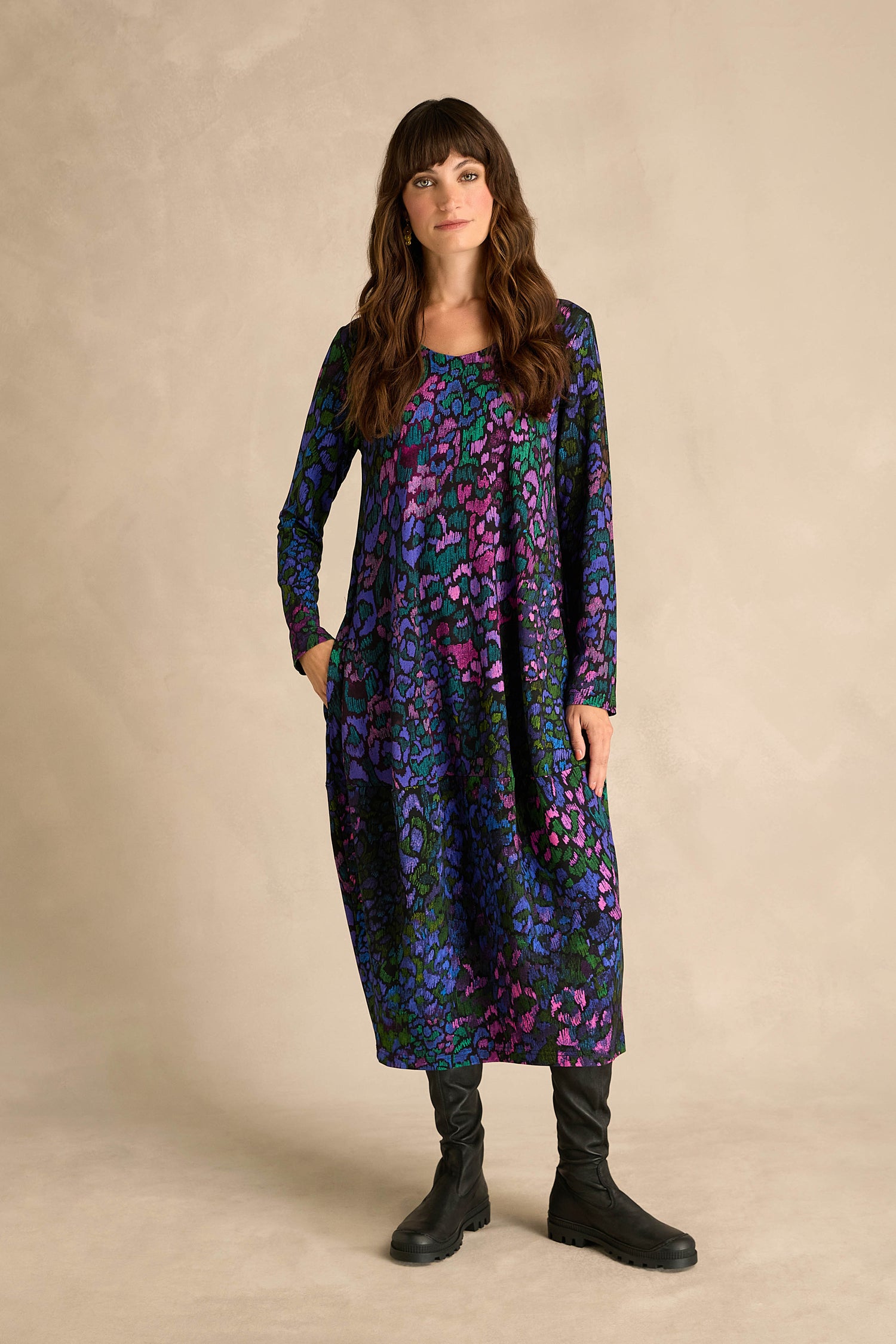 A woman poses against a plain backdrop, dressed in the Winter Garden Jersey Bubble Dress—a long-sleeve, ankle-length dress crafted from viscose jersey with an eye-catching pattern in purple, blue, and green hues. The piece features a unique bubble silhouette and is styled with black boots.