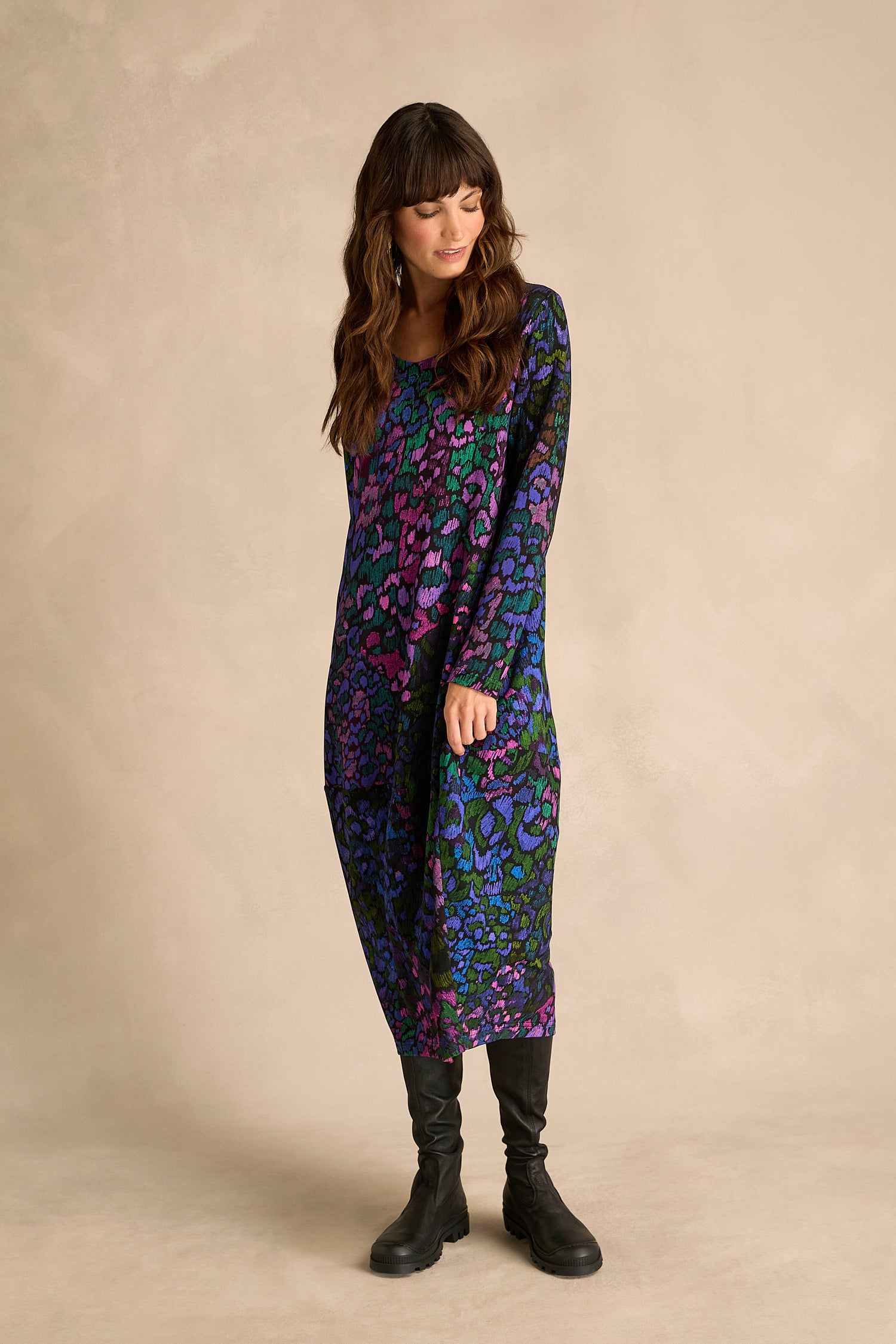 A woman wears the Winter Garden Jersey Bubble Dress, featuring a loose fit and long sleeves in soft viscose jersey with an abstract blue, purple, and green pattern. She pairs it with black boots and stands against a beige backdrop. Made in England.