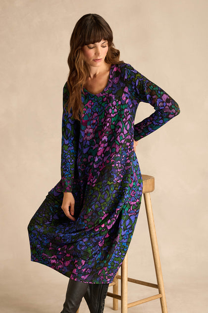 A woman wearing the Winter Garden Jersey Bubble Dress, featuring long sleeves and a vibrant blue, purple, and green pattern, sits on a wooden stool against a beige background.
