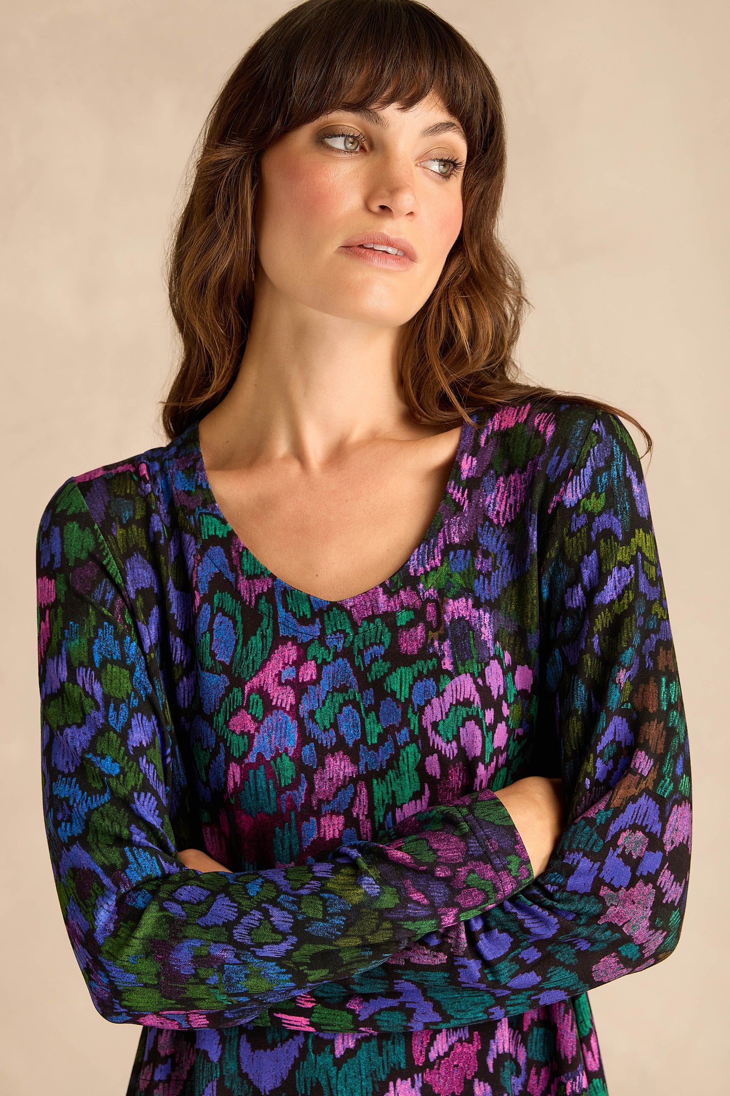 A woman with long brown hair stands against a plain background, arms crossed and looking to the side, wearing the Winter Garden Jersey Bubble Dress—a colorful, long-sleeve patterned design.