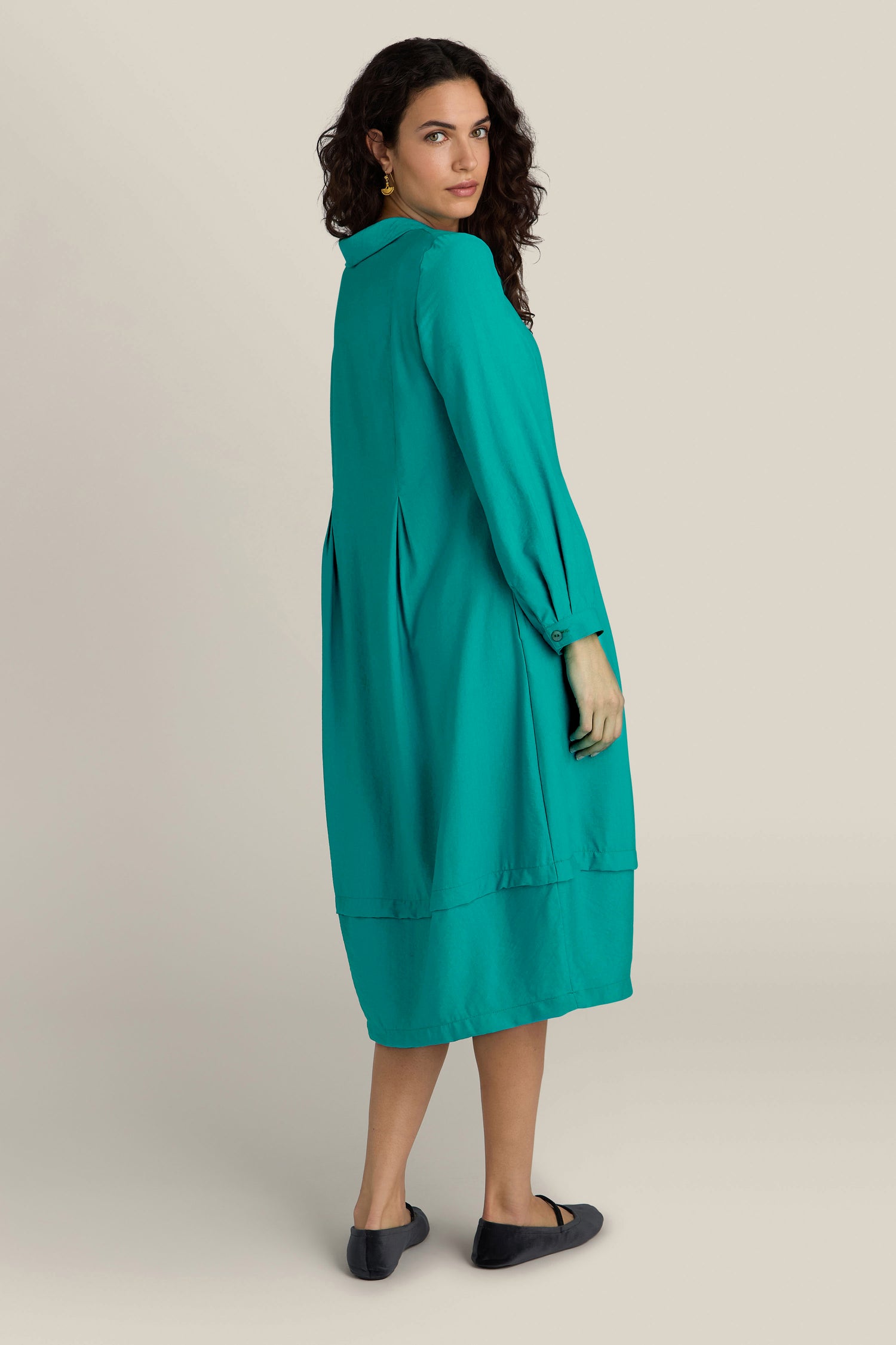 A woman with curly hair, wearing a Textured Viscose Bubble Dress in teal and black flats, radiates everyday elegance as she stands on a plain background, turned slightly to her left.