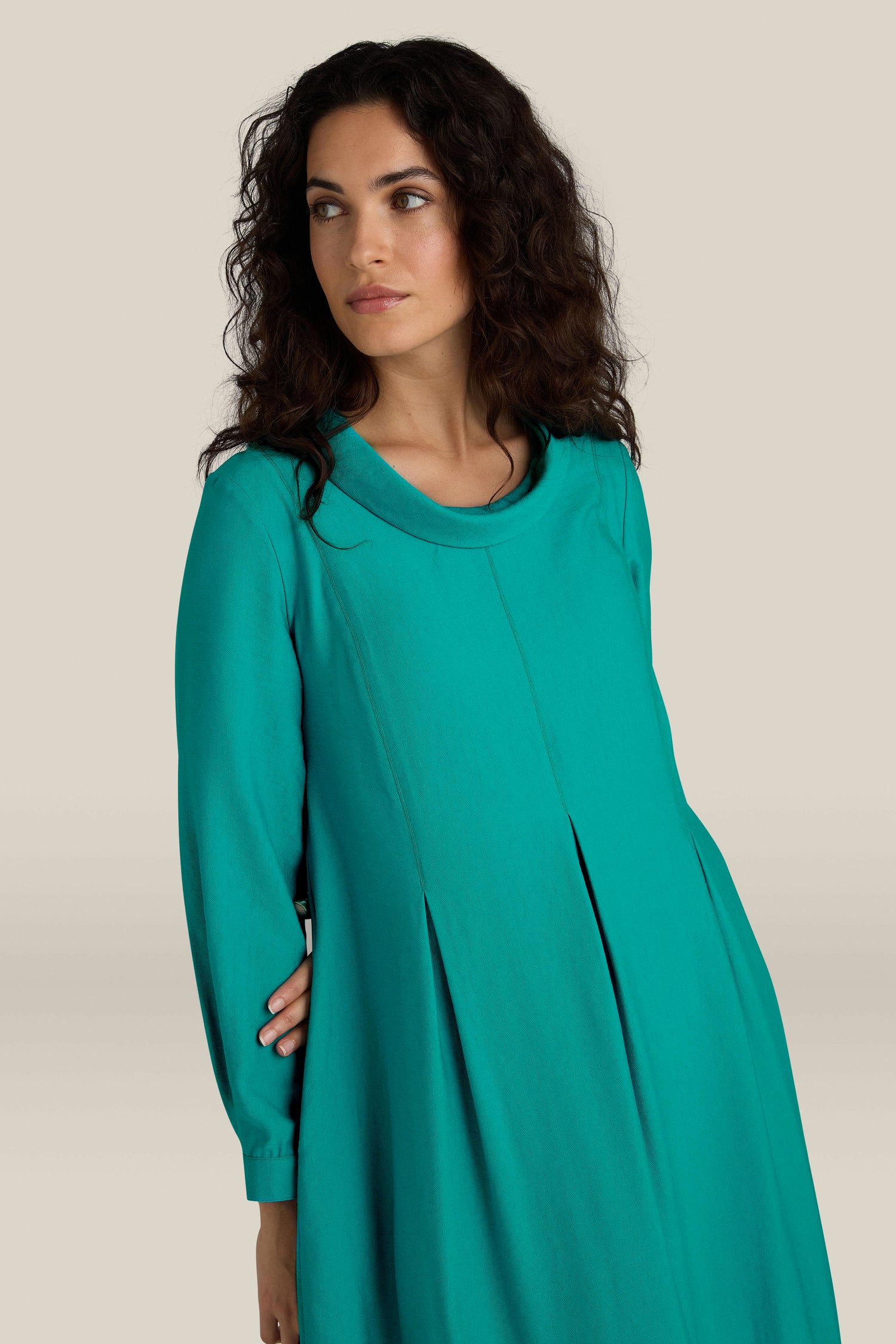 A woman with wavy dark hair, exuding everyday elegance in a long-sleeved Textured Viscose Bubble Dress in teal with button cuffs, looks to her left against a plain beige background.