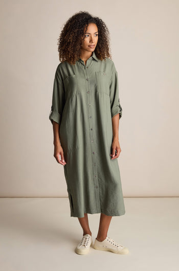 A person with curly hair stands wearing a Textured Viscose Shirt Dress in a khaki colourway and white sneakers against a plain background.