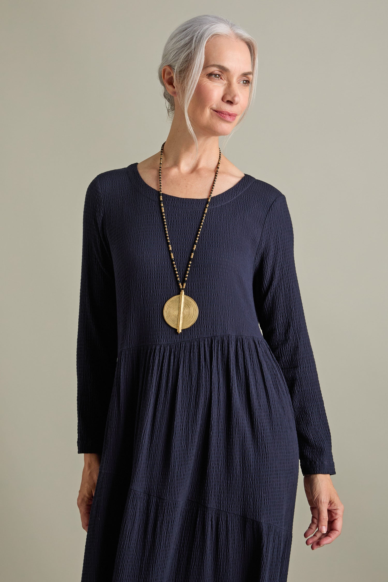 An older woman with gray hair stands against a plain background, wearing the Crinkle Viscose Tiered Dress in navy blue, featuring an elegant A-line silhouette and a maxi length. She accessorizes with a long necklace that has a large gold pendant.