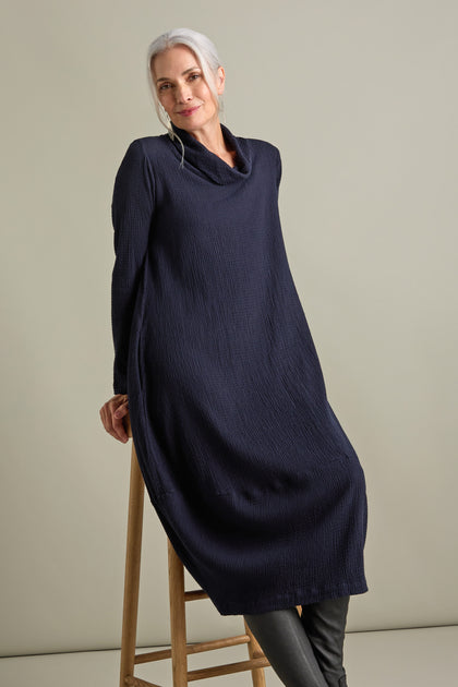 A woman with gray hair leans against a wooden stool, donning the Crinkle Viscose Bubble Dress paired with black pants. She gazes directly at the camera with a subtle smile, exuding relaxed chic against a neutral background.