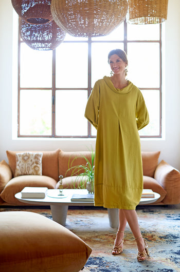 Wearing a Textured Viscose Bubble Dress, someone stands in a chic living room featuring a brown sofa, round coffee table, and large woven light fixtures. The dress's flattering fit perfectly complements the contemporary elegance of the space.