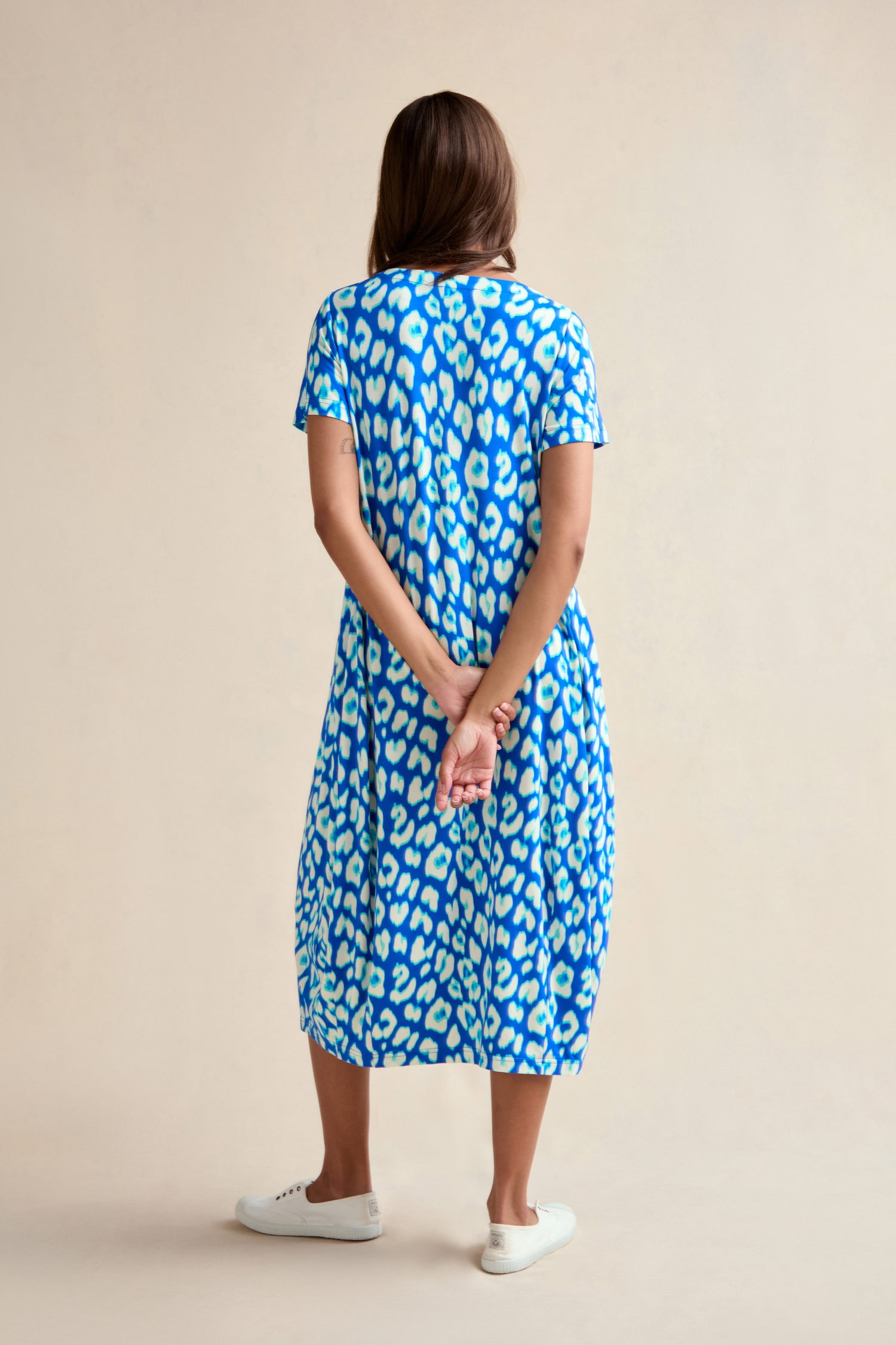 A woman stands with her back to the camera, hands clasped behind her, wearing the Leopard Print Jersey Bubble Dress and white sneakers against a plain background.