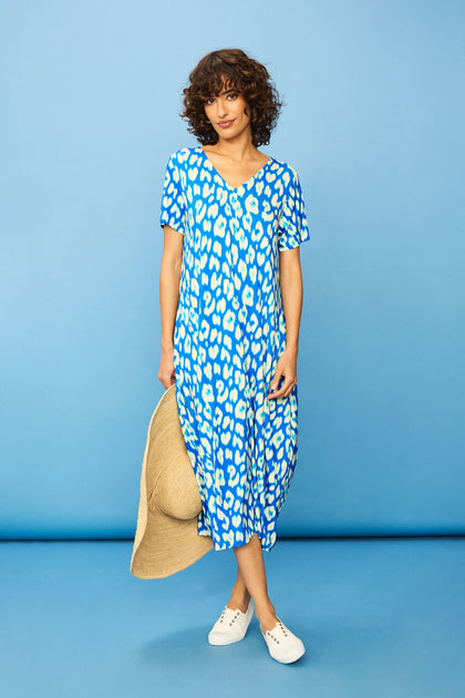 A woman stands against a solid blue background wearing the Abstract Leopard Print Jersey Bubble Dress in blue and white, holding a large floppy hat and paired with white slip-on shoes.